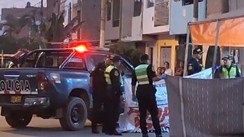 Una camioneta policial con luces intermitentes rojas en una calle, rodeada de policías y personas. Un cerco provisional bloquea parte de la escena
