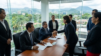 Una mujer de negocios y un hombre se dan la mano en una mesa con documentos. Varias personas observan en una oficina con vista a una ciudad y montañas.