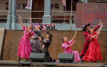 Un grupo de hombres y mujeres con vestidos rojos y trajes de gaucho baila con los brazos levantados sobre un escenario