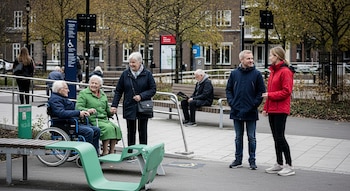 Personas de distintas edades en un parque accesible. Un hombre en silla de ruedas, dos mujeres mayores y una pareja joven interactúan. Se ven bancos verdes y rampas.