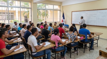Un profesor de espaldas y cerca de 30 estudiantes jóvenes sentados en pupitres en un aula. Hay una pizarra blanca con texto y una bandera dominicana.