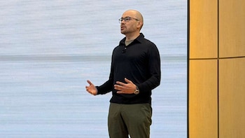 Demis Hassabis, un hombre calvo con gafas, camisa oscura y pantalones verdes, gesticula frente a una pantalla y un panel de madera