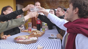 Brazos de una persona utilizando pinzas y espátula sobre parrilla con carne, chorizos y pan; al fondo, jóvenes sentados alrededor de una mesa