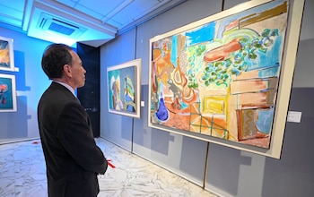Un hombre en traje oscuro de perfil observa un cuadro abstracto grande en una galería. Se ven otras pinturas, paredes grises, suelo de mármol y luz azul