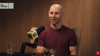 Adam Grant frente a un micrófono, un vaso de licor y un recipiente con piedras sobre una mesa ante una pared de madera