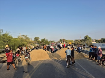 Una carretera bloqueada por dos grandes montículos de arena, con grupos de personas a ambos lados y motocicletas. El cielo es azul claro y se ven árboles al fondo