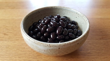 Un bol de cerámica de color beige con motas marrones contiene porotos negros cocidos. El bol está apoyado sobre una superficie de madera clara.