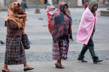 Personas con abrigos, bufandas, gorros y paraguas caminan por una calle húmeda