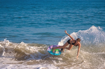 Una mujer de cabello castaño y bikini negro sobre una tabla de skimboard colorida sobre una ola en la playa, con el mar azul de fondo