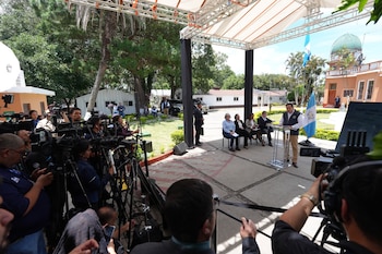 Hombre en un podio transparente habla en conferencia de prensa al aire libre. Periodistas con cámaras y equipos observan. Bandera de Guatemala visible