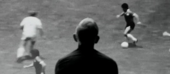 Imagen en blanco y negro granulada de un campo de fútbol. Un jugador de espaldas en primer plano, y dos jugadores más, uno con el balón, en el fondo