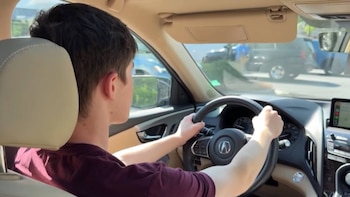 Adolescente con camiseta granate conduce un coche. Sus manos sujetan el volante de un Acura. Interior beige del vehículo. Vehículos al fondo