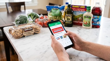 Manos sosteniendo un teléfono móvil con la interfaz de Uber Eats y Costco frente a empaques de galletas, vegetales y botellas sobre una mesa.