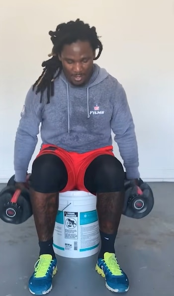 Hombre con sudadera gris y pantalones cortos rojos sentado en un cubo blanco mientras sostiene dos mancuernas pesadas