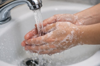 Manos con espuma de jabón bajo el chorro de agua de un grifo metálico en un lavamanos blanco, con salpicaduras de agua.