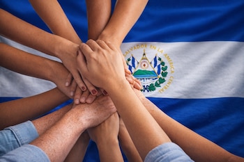 Primer plano de múltiples manos de personas apiladas, sobre la bandera de El Salvador que muestra dos franjas azules, una blanca central y su escudo.