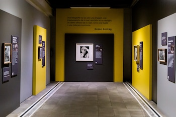 Sala de exposición con fotografías enmarcadas en paredes grises y amarillas, cédulas explicativas y una cita en la pared del fondo