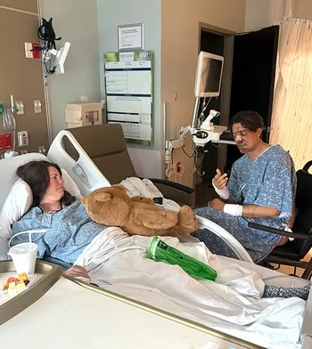 Una mujer recostada en una cama de hospital con un oso de peluche habla con un hombre lesionado sentado en una silla a su lado