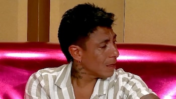 Brian Sarmiento, un hombre de cabello oscuro y camisa a rayas claras, sentado en un sofá magenta, con lágrimas visibles en sus ojos mientras mira hacia abajo