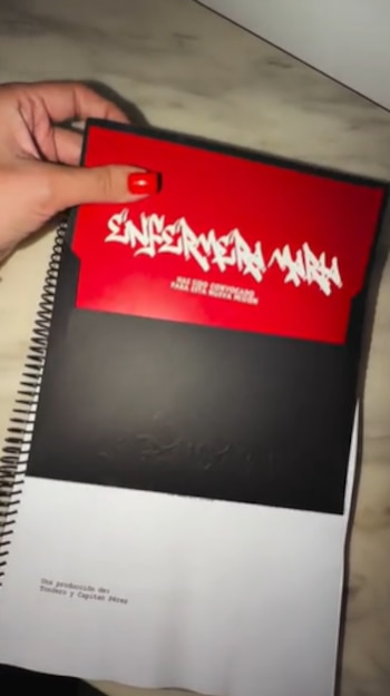 Mano de mujer con uñas rojas sosteniendo un guion anillado de tapa negra. Se ve una tarjeta roja con el texto blanco "ENFERMERA MALA"