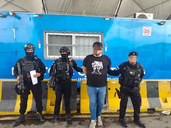 Tres agentes de seguridad custodian a un hombre esposado frente a un contenedor azul y barreras amarillas