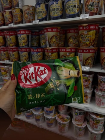Una mano con esmalte de uñas rojo sostiene un paquete de Kit Kat verde sabor matcha en un supermercado con fideos instantáneos al fondo