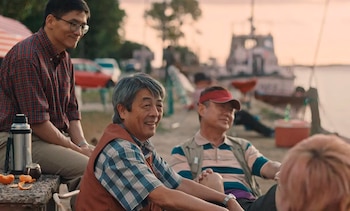 Primer plano de tres hombres asiáticos sonriendo y sentados al aire libre; en primer plano hay un termo, un mate y cáscaras de naranja; un barco se ve borroso al fondo