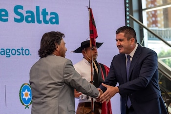 Dos hombres con traje se estrechan la mano frente a una pantalla con la palabra Salta y un hombre con atuendo tradicional que sostiene una bandera