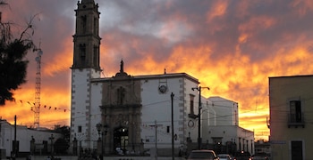 Edificio de iglesia blanca con campanario y fachada ornamentada, un cielo de atardecer anaranjado y rojizo. Se ven farolas, coches y personas
