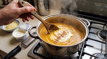 Una mano sostiene una cuchara de madera en una olla con sopa de zapallo sobre la cocina, junto a un cuenco con queso y un rallador.