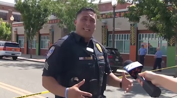Un oficial de policía uniformado de pie en una calle, con su mano gesticulando mientras habla a un micrófono sostenido por una persona; un edificio y vehículos al fondo