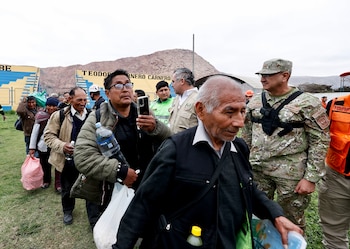 Grupo de personas con bolsas y mochilas camina en fila en un estadio junto a militares y personal de defensa civil