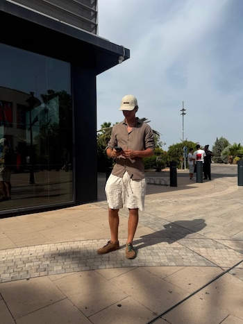 Hombre joven con gorra, camisa y pantalones cortos de color claro mirando un teléfono. Se encuentra en una zona pavimentada con un edificio y vegetación