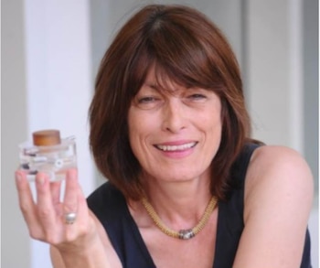 Mujer de mediana edad con cabello castaño sonríe y sostiene un pequeño dispositivo transparente. Viste una camiseta oscura y un collar dorado con un abalorio
