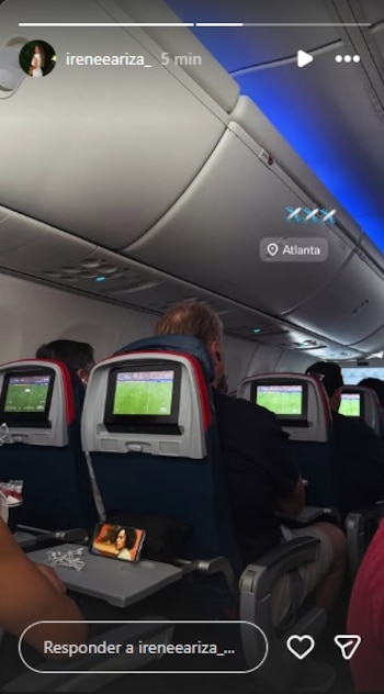 Cabina de avión con pasajeros. Pantallas de entretenimiento con partido de fútbol. Teléfono móvil sobre mesa. Interfaz de red social: "ireneeariza_", "Atlanta"