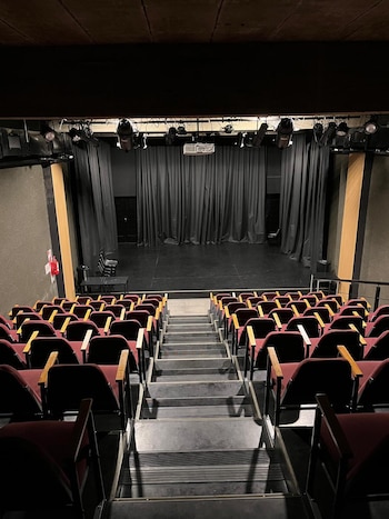 Interior de un teatro pequeño con butacas vacías de color vino tinto, escaleras centrales y un escenario oscuro con telones negros y luces superiores
