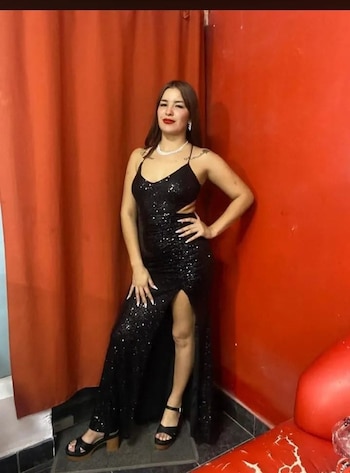 Una mujer joven con un vestido de lentejuelas negro, un collar de perlas, pendientes y sandalias de plataforma. Fondo de cortina naranja y pared roja