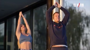Un hombre y una mujer realizan una pose de yoga con los brazos elevados y las manos unidas. El hombre viste una sudadera oscura, la mujer un top deportivo azul. Se ve un logo "EL A/T19"