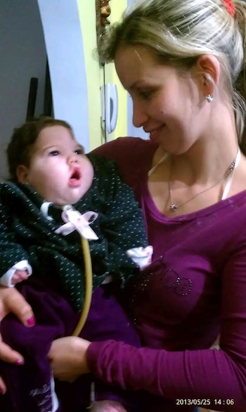 Mujer rubia con camisa morada sostiene a un bebé con cabello oscuro y un tubo de alimentación visible