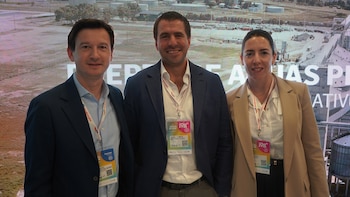 Dos hombres y una mujer con sacos formales y credenciales colgadas posan juntos delante de una pantalla digital con imágenes