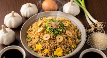 Plato de arroz frito con trozos de ajo, huevo revuelto, cebolletas y semillas de sésamo humeante; rodeado de ajos enteros, cebollas verdes, huevo, arroz y salsa de soja.