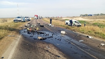 Una carretera rural con restos de un accidente, dos vehículos blancos dañados, personal de seguridad y una fila de camiones en el fondo