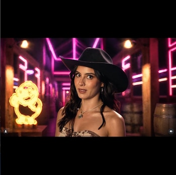 Mujer con cabello oscuro, sombrero vaquero negro, top strapless, collar y aretes; fondo oscuro con luces de neón fucsia, figura luminosa amarilla y dos barriles de madera