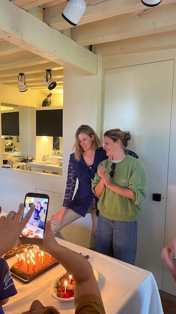 Bernardita Barreiro y Agustina Macri sonríen en una habitación con techo de vigas. En primer plano, pasteles con velas y una persona grabando con un móvil