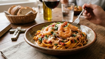 Primer plano de un plato de pasta con camarones, salsa rosada, queso rallado y cilantro, sobre una mesa de madera. Una mano sostiene un tenedor