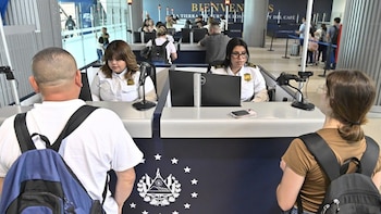 Vista trasera de dos viajeros con mochilas frente a agentes de migración en El Salvador, sentados en sus puestos con computadoras en un aeropuerto