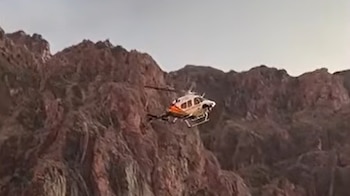 Un helicóptero de color blanco y anaranjado vuela cerca de laderas rocosas y empinadas en un cañón