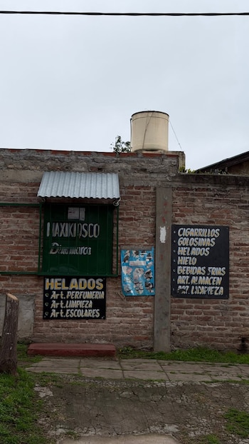 Muro de ladrillos, cartel verde "MAXIKIOSCO", toldo metálico corrugado, dos carteles negros con texto, cartel azul, poste de concreto, tanque de agua, vereda, pasto, cable