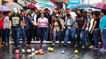 Personas con paraguas observan a varios participantes que lanzan melones y pelotas sobre el asfalto mojado de una calle.