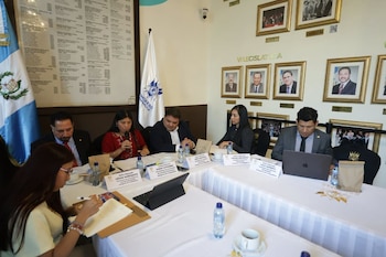 Varias personas sentadas alrededor de una mesa larga con banderas, cuadros en la pared, tarjetas de identificación, micrófonos, portátiles y botellas de agua